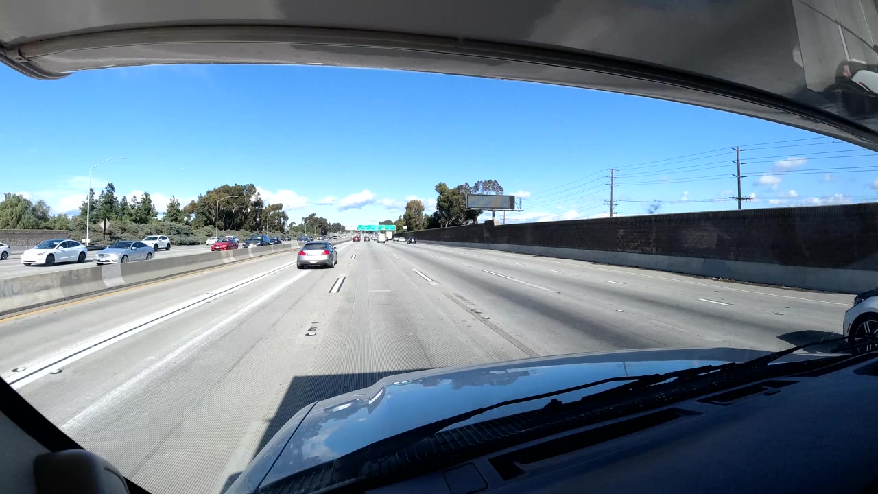 Forward dashboard view on a California coast freeway with cars in multiple lanes, a sound wall, distant road signs, power lines, and no visible people.