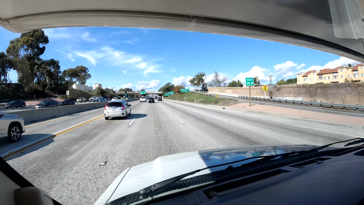 Central Coast California freeway view from a vehicle with cars ahead, trees on the roadside, a green exit sign in the distance, bright blue sky, and no visible people.