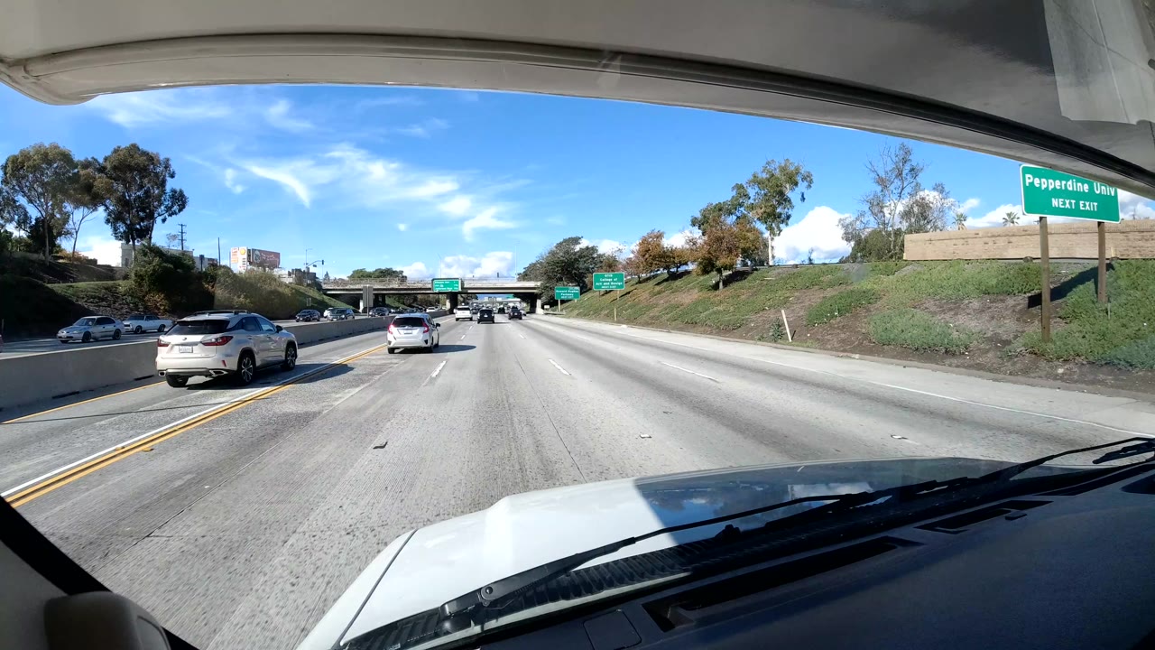 Central Coast California road POV with several vehicles ahead, a grassy embankment, roadside trees, distant highway signage, the vehicle hood at the bottom, and no visible people.