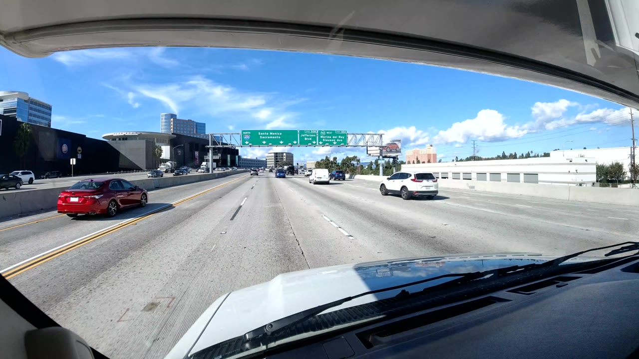Central Coast California freeway scene with a red car at left, white SUV at right, overhead green signs, an overpass structure, sound wall, blue sky, and no visible people.