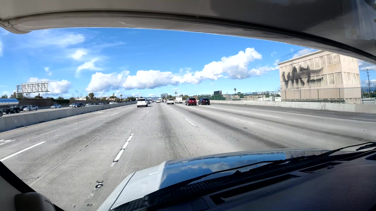 Forward-facing California coast freeway view from inside a vehicle, with the hood at the bottom, cars ahead, concrete barriers, a large building at right, and no visible people.