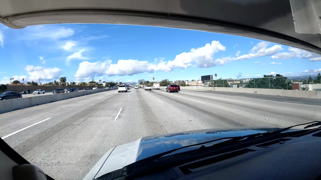 Wide highway lanes with moving cars and clouds viewed through a windshield.