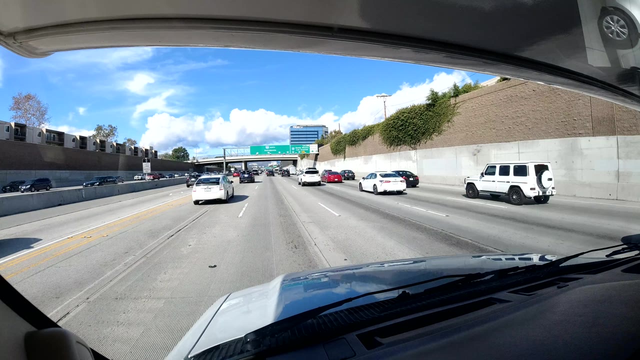 California coast freeway traffic passes concrete retaining walls, roadside greenery, overhead green signs, and urban buildings, with the vehicle hood visible and no visible people.