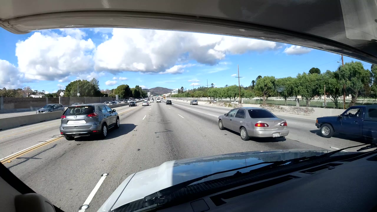 Forward windshield view of highway traffic with cars, trees, clouds, and vehicle hood visible.