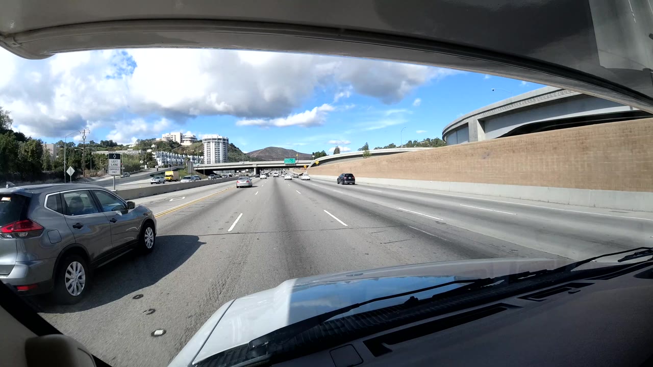 California coast highway dashcam frame showing a car passing near the camera, concrete sound wall, overpass structure, distant buildings, clear blue sky, and no visible people.