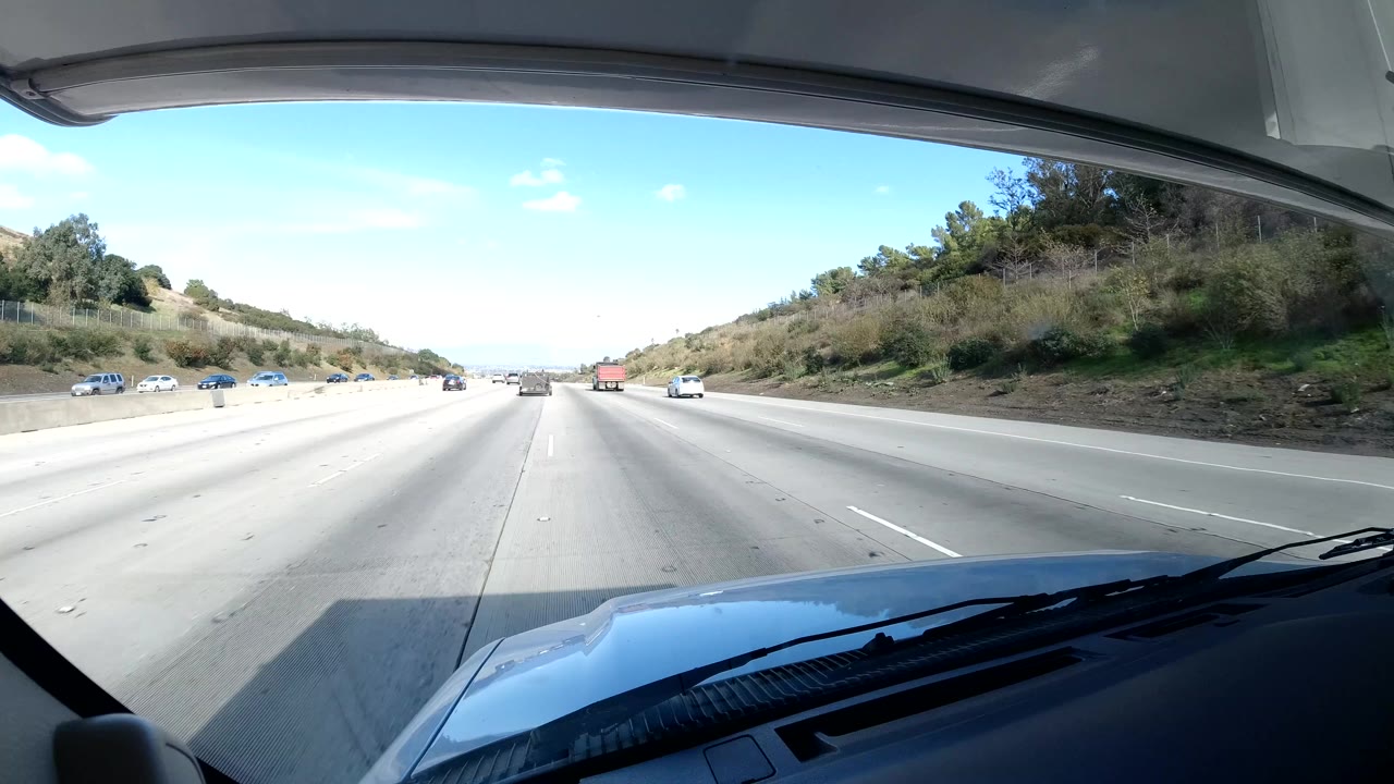 Coast California highway view from inside a vehicle, with open lanes, distant cars, roadside greenery, blue sky, dashboard foreground, and no visible people.