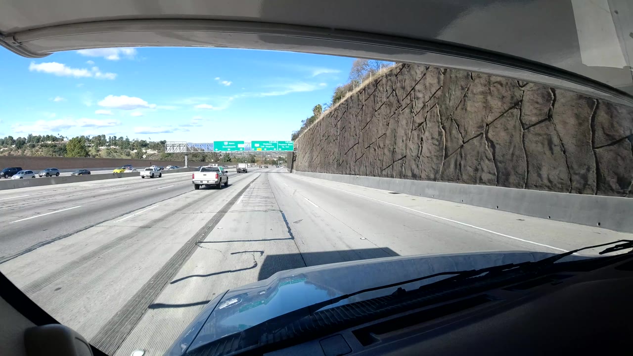 Coast California roadway viewed through a windshield beside a textured retaining wall, with cars, overhead green signs, road shadows, dashboard foreground, and no visible people.