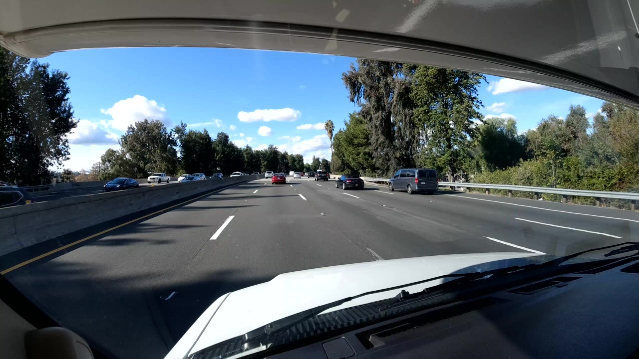 Forward in-vehicle view on a Coast California freeway with the white hood and windshield wiper visible, cars ahead, roadside trees, blue sky, and no visible people.
