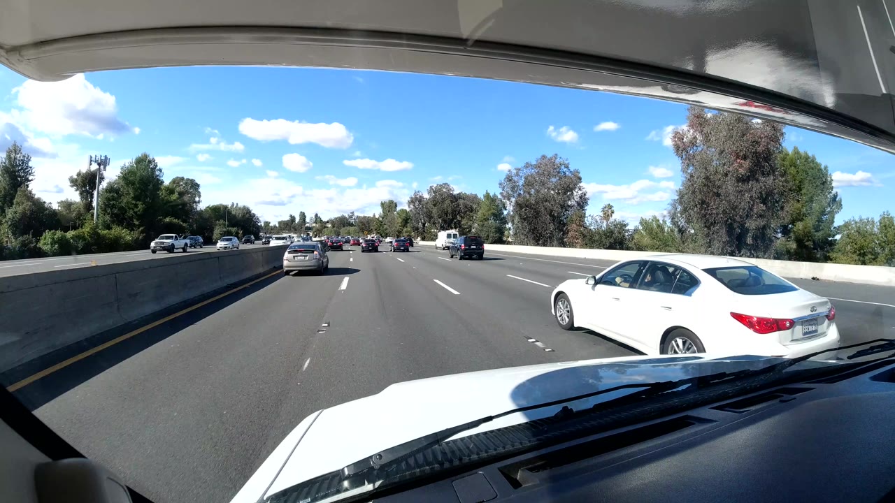 The Coast California freeway view opens wider to the right lane, where a white sedan passes near the camera, with trees, a concrete barrier, blue sky, and no visible people.