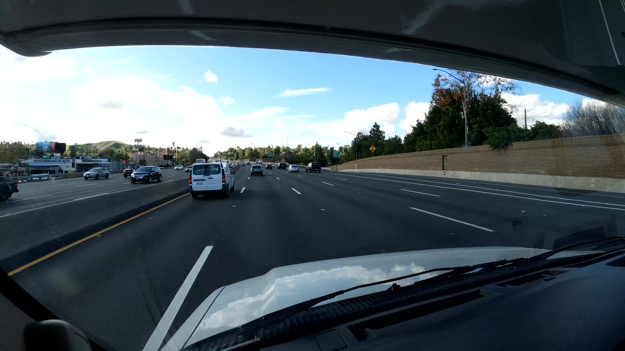 Dashcam view on the California coast shows multi-lane freeway traffic ahead, a white vehicle hood at the bottom edge, roadside wall and trees, bright sky, and no visible people.