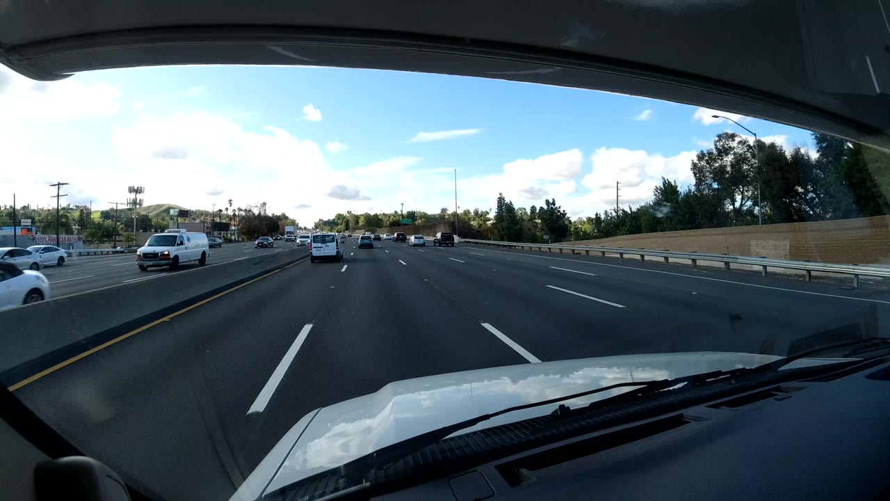A California coast freeway scene is viewed from inside the vehicle, with lane markings, cars and a van in midground, trees along the right side, distant hills, and no visible people.