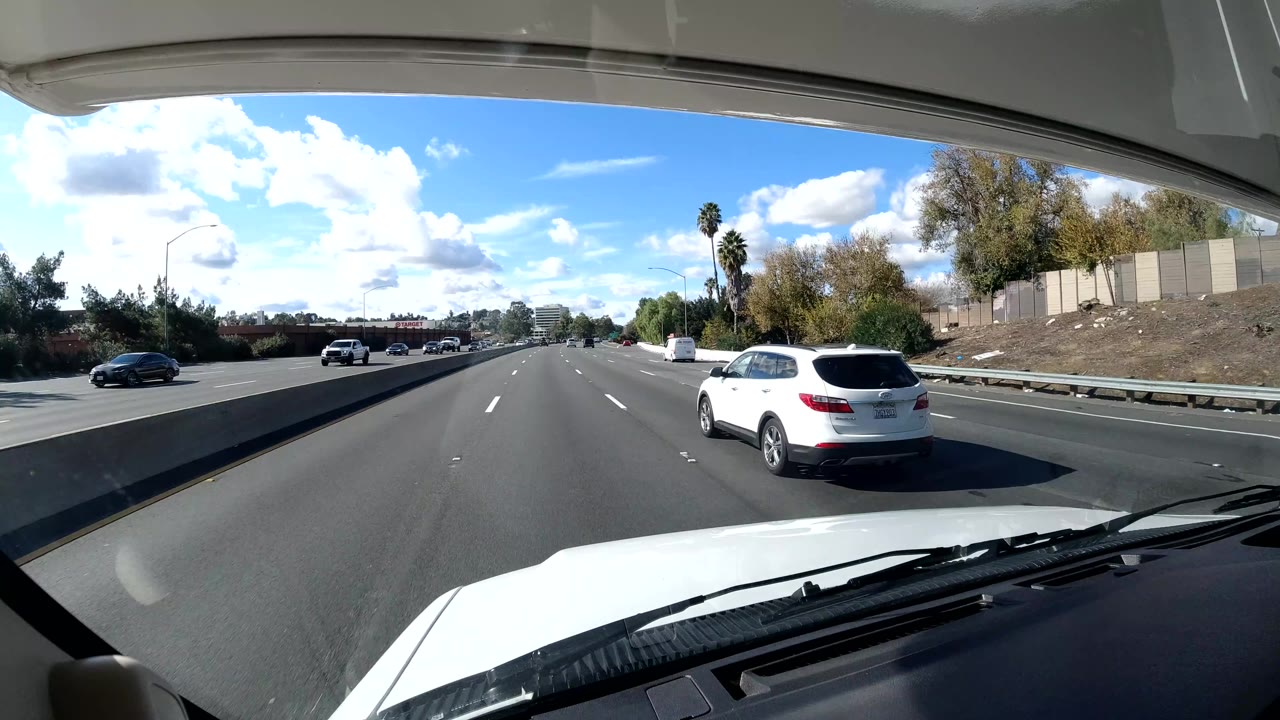 The vehicle continues along a California coast freeway, with a white SUV near the right lane, guardrail and trees at roadside, open lanes ahead, bright sun, and no visible people.