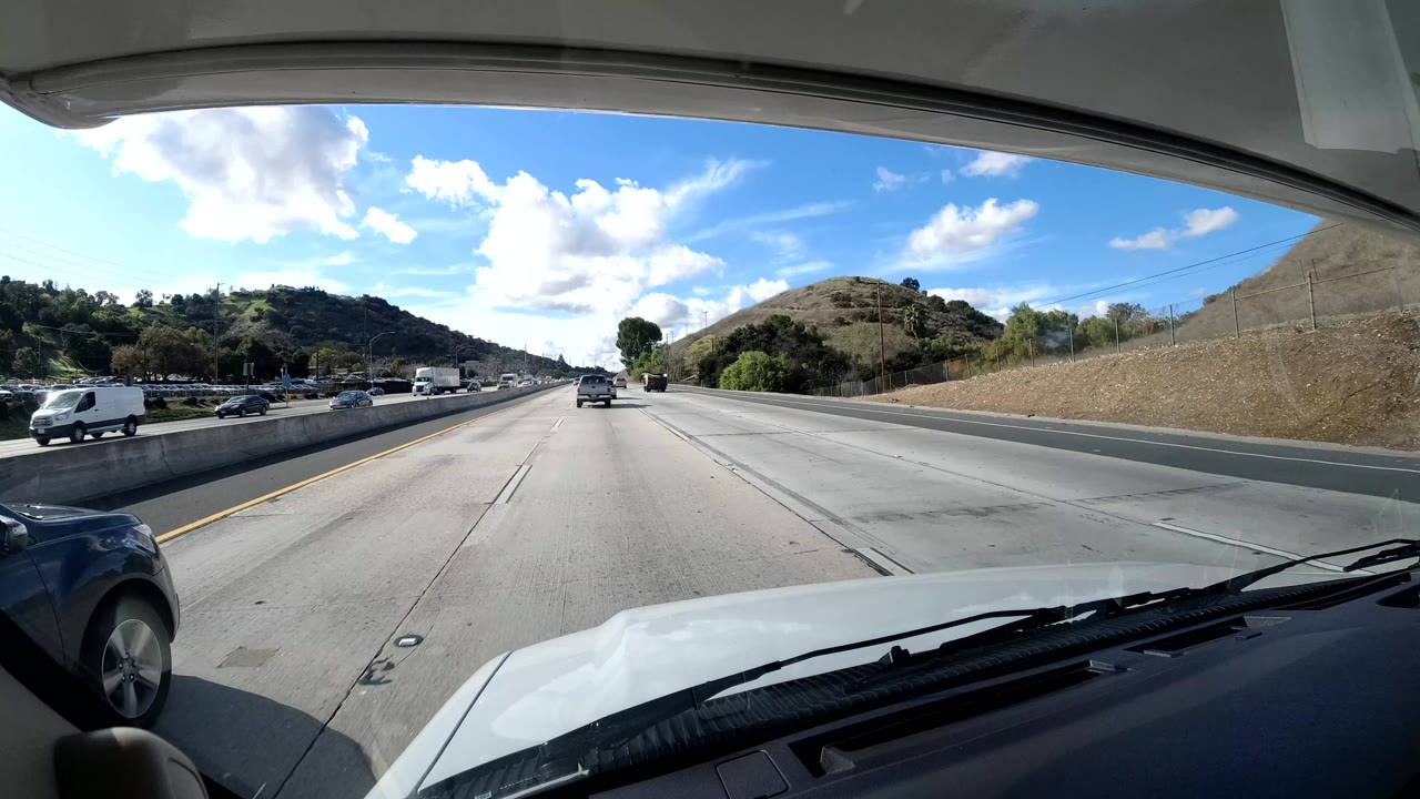 Central Coast California freeway view from inside a vehicle, showing dashboard foreground, multiple lanes, dry hills, cars, trucks, and no visible people.