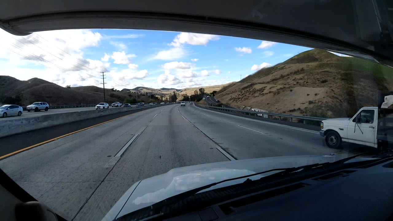 Central Coast California freeway from a windshield viewpoint, with a white truck at the right edge, cars across the lanes, hillside terrain, and no visible people.