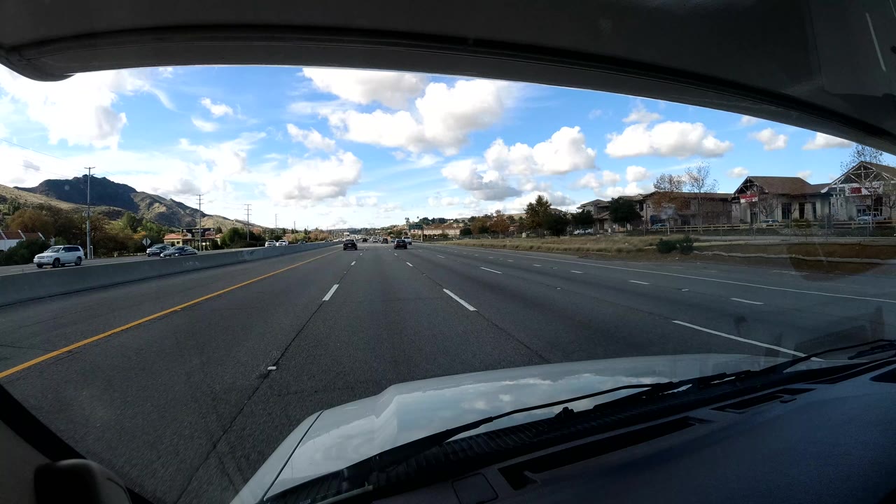 Forward windshield view on a California coast highway with sparse traffic ahead, hillside homes to the right, mountains in the distance, bright clouds overhead, and no visible people.