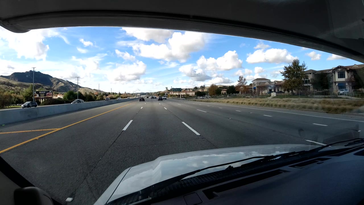 California coast freeway traffic continues from a dashboard viewpoint, with cars ahead in the center lanes, hillside houses on the right shoulder area, distant hills, and no visible people.