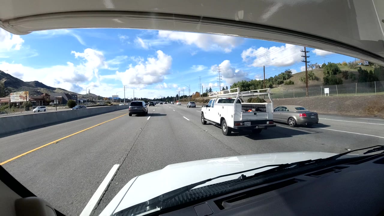 A California coast highway scene shows a white pickup in the right lane, cars ahead, a sound wall and utility poles along the roadside, hills beyond, and no visible people.