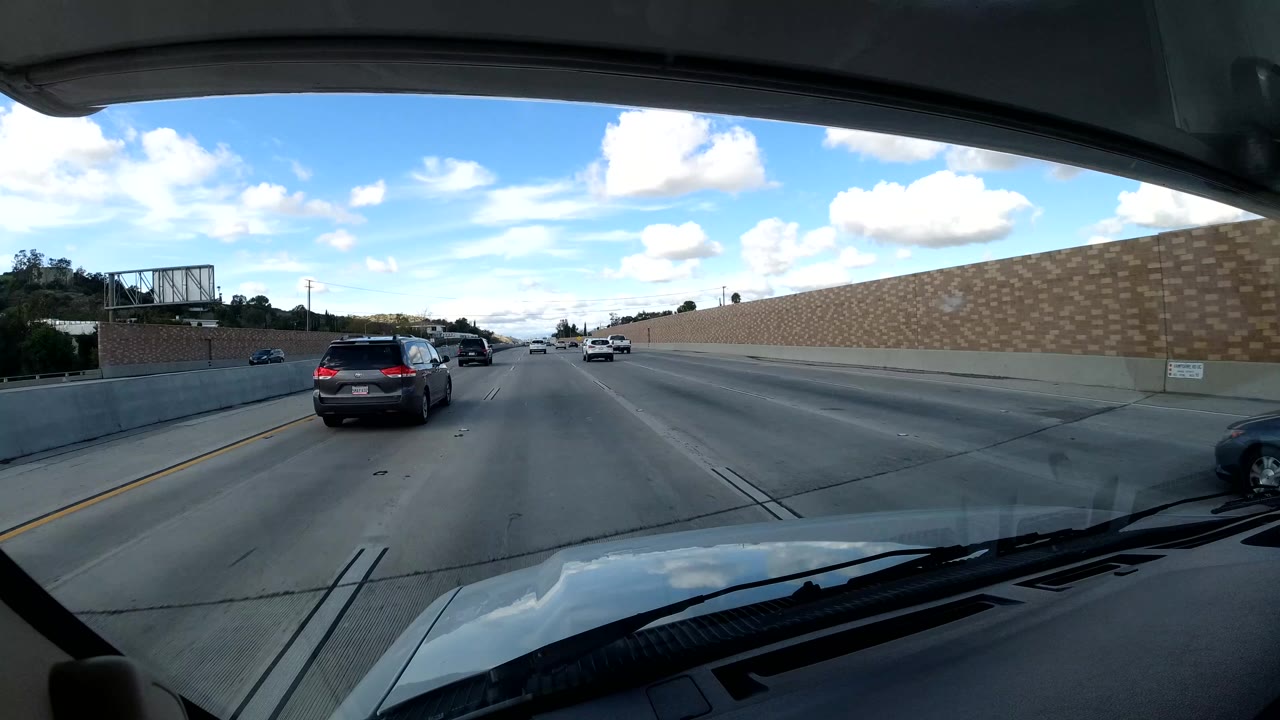 California coast freeway lanes seen from inside a vehicle, with cars ahead, a concrete sound wall, windshield frame, and no visible people.