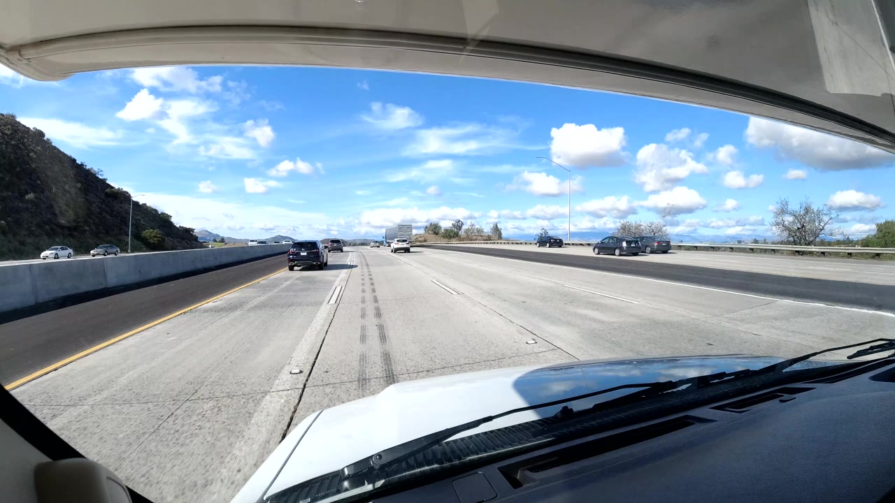 California coast freeway scene from a front vehicle angle, with open lanes, cars ahead, a hillside at left, bright sky, and no visible people.