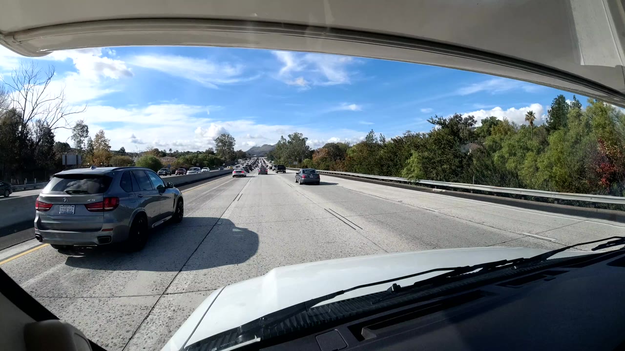 A California coast freeway view from inside a vehicle shows a gray SUV in the left lane, trees along the road, blue sky, and no visible people.