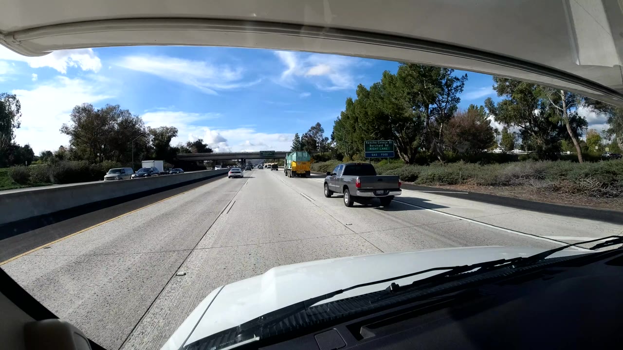The California coast highway view shows pickup trucks ahead, trees on the right shoulder, a green road sign in the distance, and no visible people.