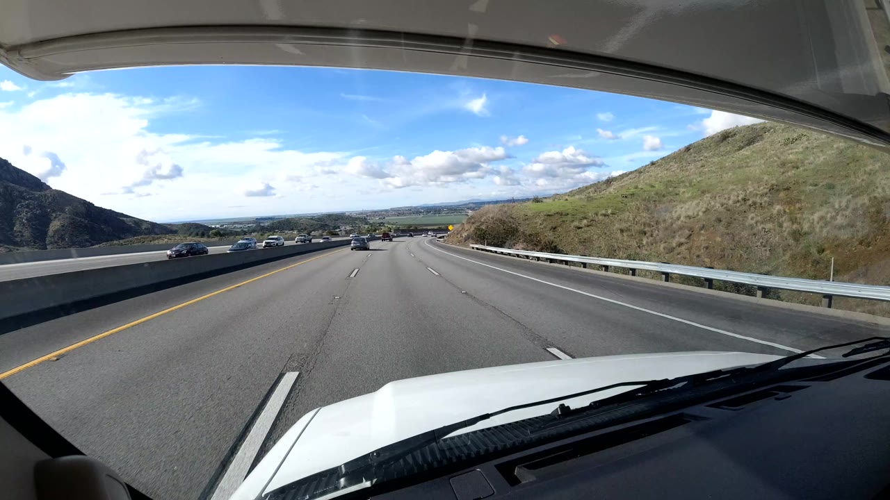 Dashcam-style view from a vehicle on a Coast California highway, with the white hood at the bottom, cars ahead, hills on both sides, scattered clouds, and no visible people.