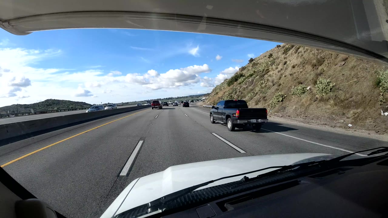 Dashcam view on a Coast California highway with a dark pickup in the right lane, several cars ahead, a rocky hillside beside the road, bright clouds, and no visible people.
