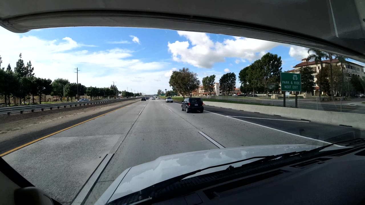 California coast highway view with cars spread across open lanes, a green roadside sign on the right, trees and buildings behind a sound wall, and no visible people.