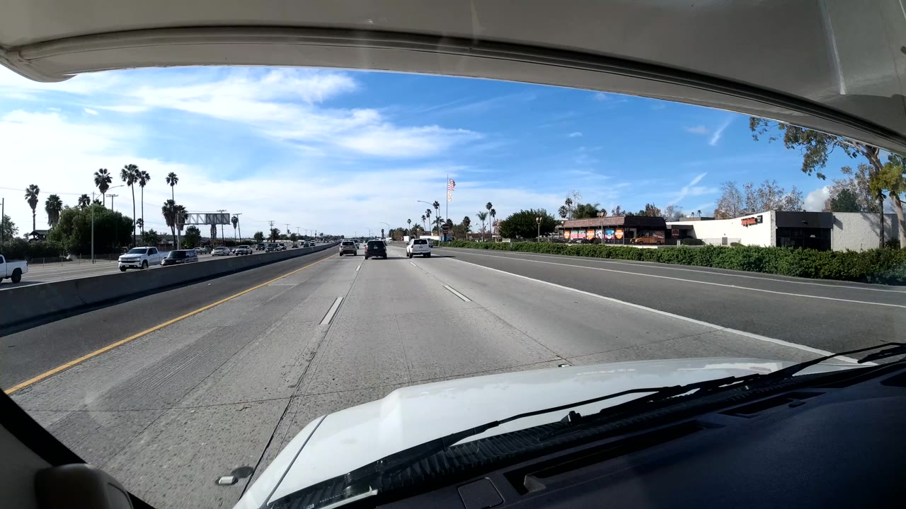 The freeway opens ahead in Central Coast California, with several cars in the lanes, roadside commercial buildings to the right, palm trees, blue sky, and no visible people.