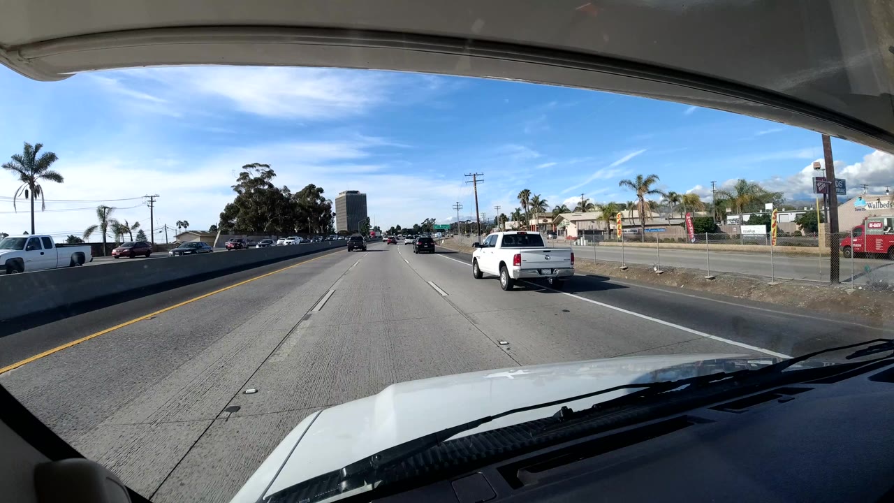 Forward-facing Coast California freeway view from a vehicle, with the hood at the bottom, cars ahead, roadside trees and buildings, blue sky, and no visible people.
