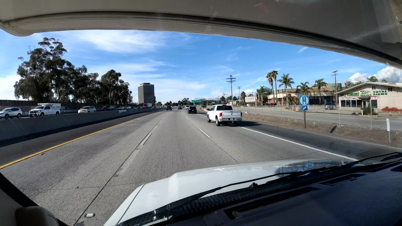Coast California freeway frame with a white pickup ahead, roadside businesses to the right, a blue transit-style sign, trees, and no visible people.