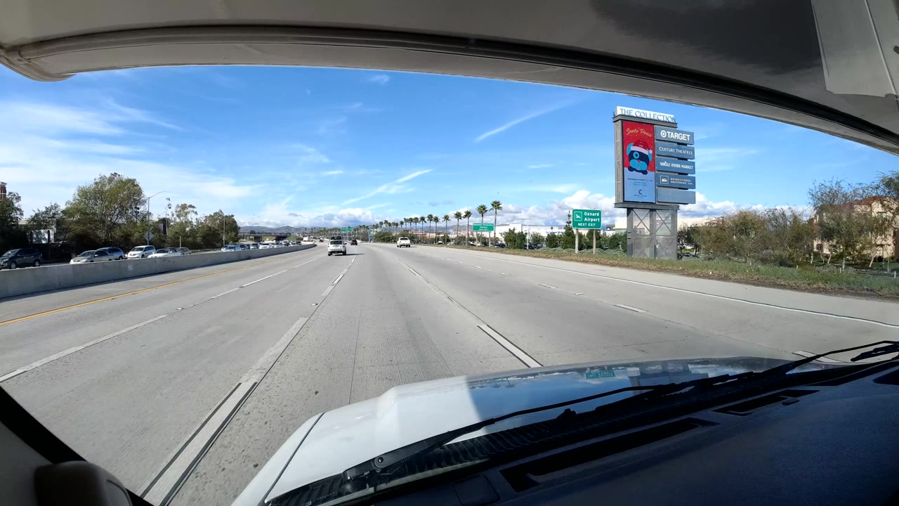 Wide Coast California freeway view with open lanes, a tall roadside sign tower, distant mountains, sparse cars, blue sky, and no visible people.