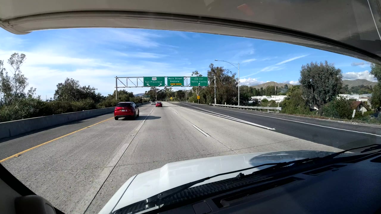 Central Coast California dashboard-view freeway frame with the vehicle hood at bottom, overhead green road signs, several cars ahead, trees, hills, blue sky, and no visible people.