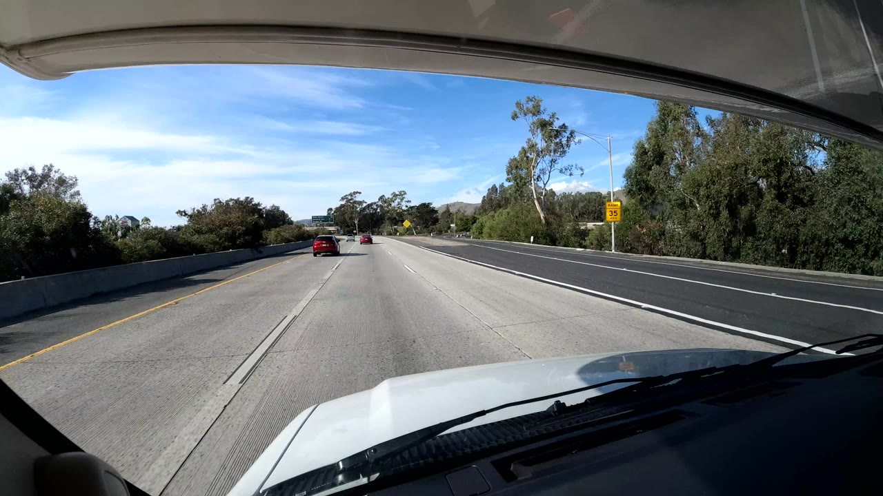Central Coast California freeway view from the dashboard, with the hood in the foreground, cars in the distance, roadside trees and shrubs, hills beyond the lanes, and no visible people.