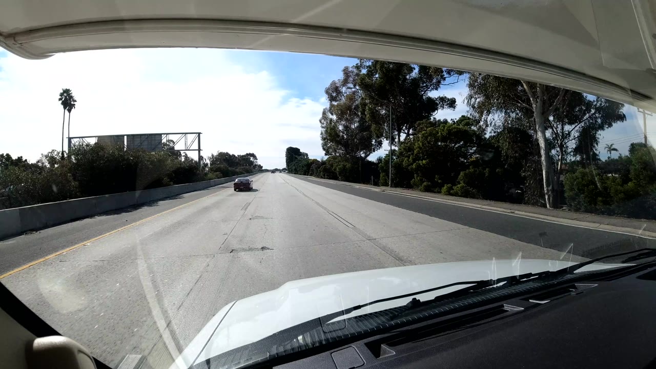 Central Coast California dashboard freeway frame with bright pavement, a sign gantry ahead, tall trees and a palm at the roadside, the vehicle hood at bottom, and no visible people.