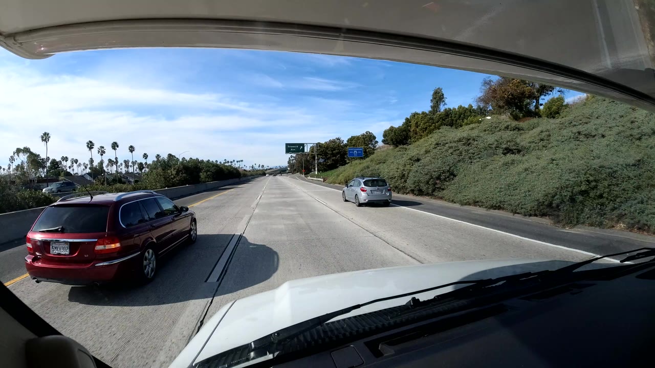 Dashboard freeway view with a red minivan passing on the left and palms in the distance.