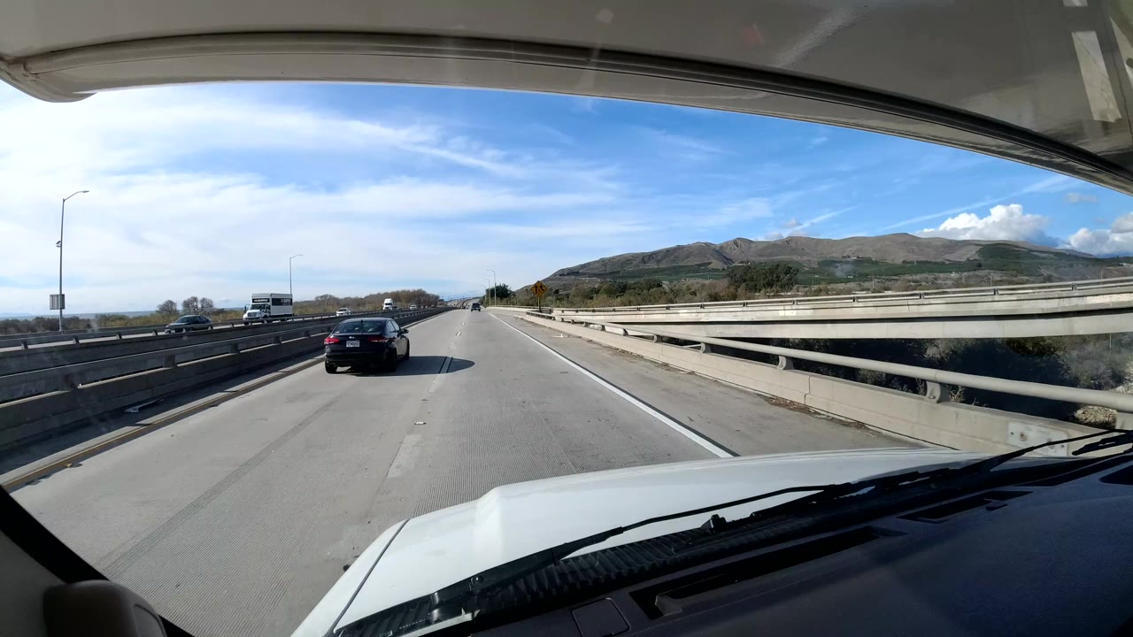 Forward view from inside a vehicle on a California coast highway, with the vehicle hood, windshield edge, bridge railings, cars ahead, low hills, blue sky, and no visible people.