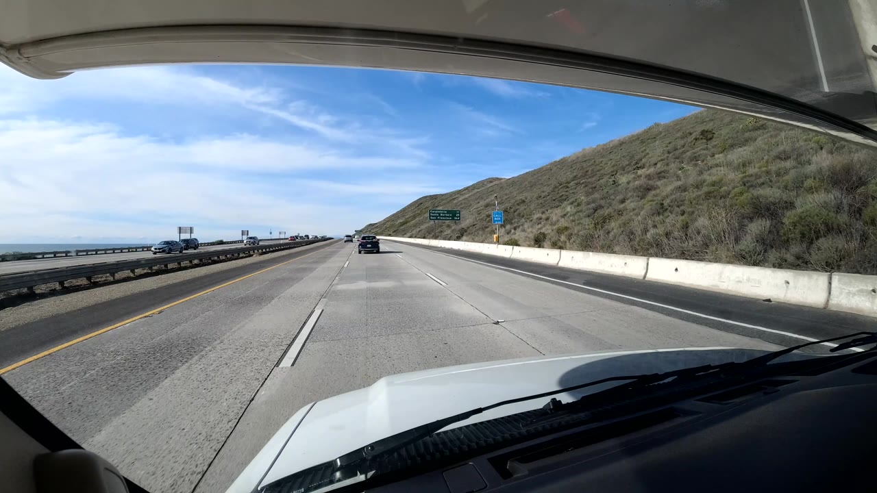 California coast highway view from behind a windshield, showing the vehicle hood, broad concrete lanes, hillside embankment, roadside signs, scattered vehicles, blue sky, and no visible people.
