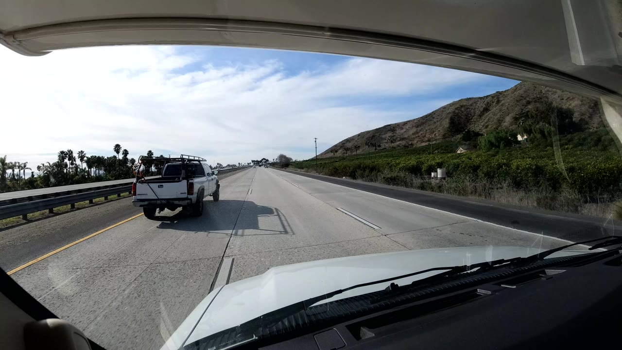 Forward windshield view on a California coast highway with a white pickup ahead, two people visible in the truck bed, dry hillside on the right, and bright sky overhead.