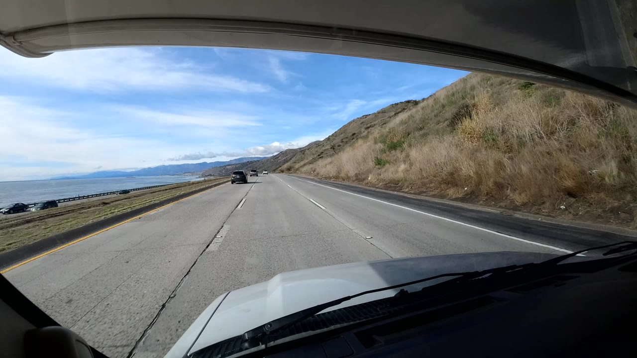 California coast highway view through the windshield with ocean water and distant mountains on the left, dry hillside on the right, several cars ahead, and no visible people.