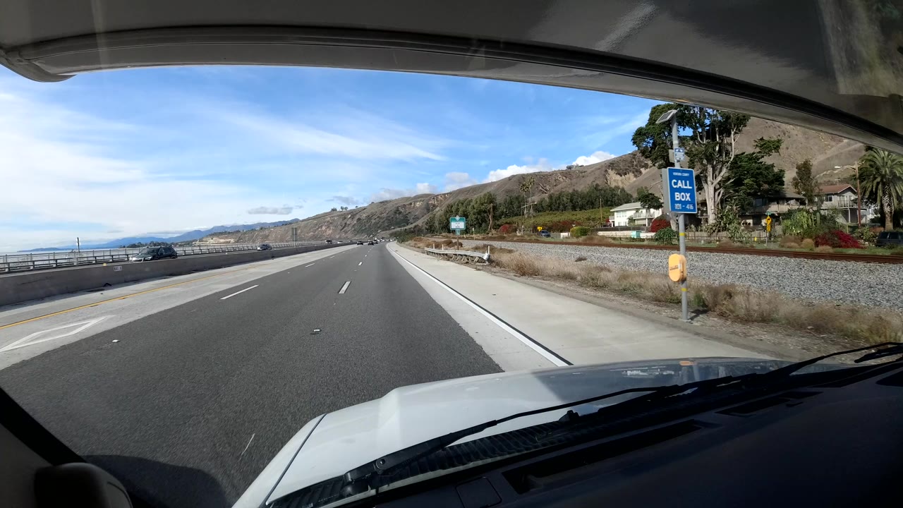 Forward vehicle view on a Coast California highway with a white hood in the foreground, a roadside call box sign, hills, and no visible people.