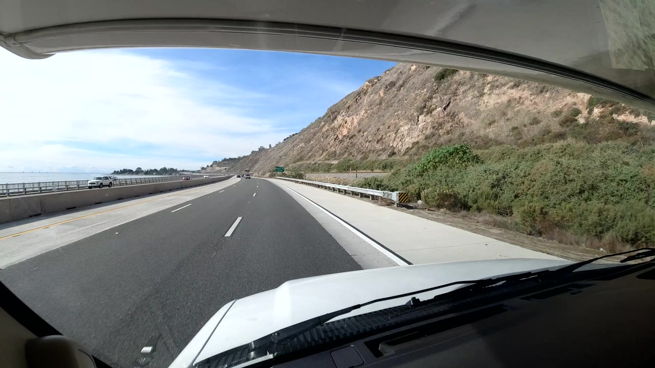 Coast California highway lane beside a steep tan hillside, green roadside vegetation, concrete barrier, distant cars, and no visible people.