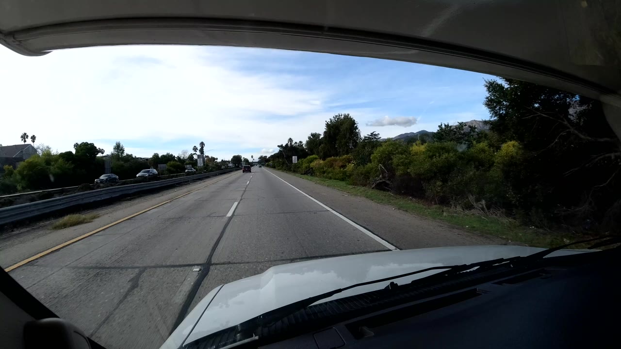 Forward windshield view on a Coast California road with a white vehicle hood, lane markings, roadside trees, a guardrail, distant hills, and no visible people.