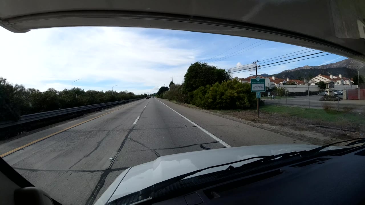The vehicle passes a more developed Coast California roadside, with lanes ahead, a sign on the right, homes or low buildings, utility lines, hills, and no visible people.