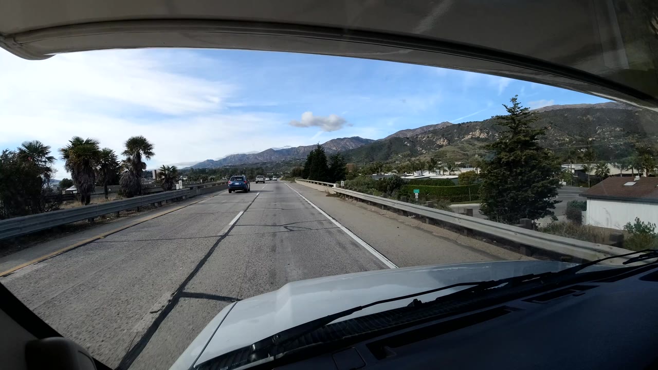 Vehicle POV on highway with palms, guardrail, and hills