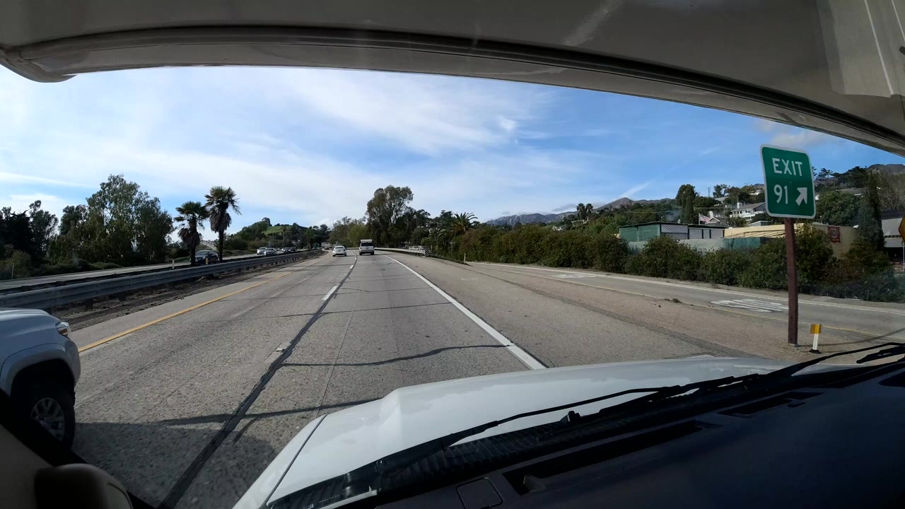 Forward-facing view from a vehicle on a coastal California highway, with an exit sign on the right shoulder, cars ahead, green trees, and no visible people.