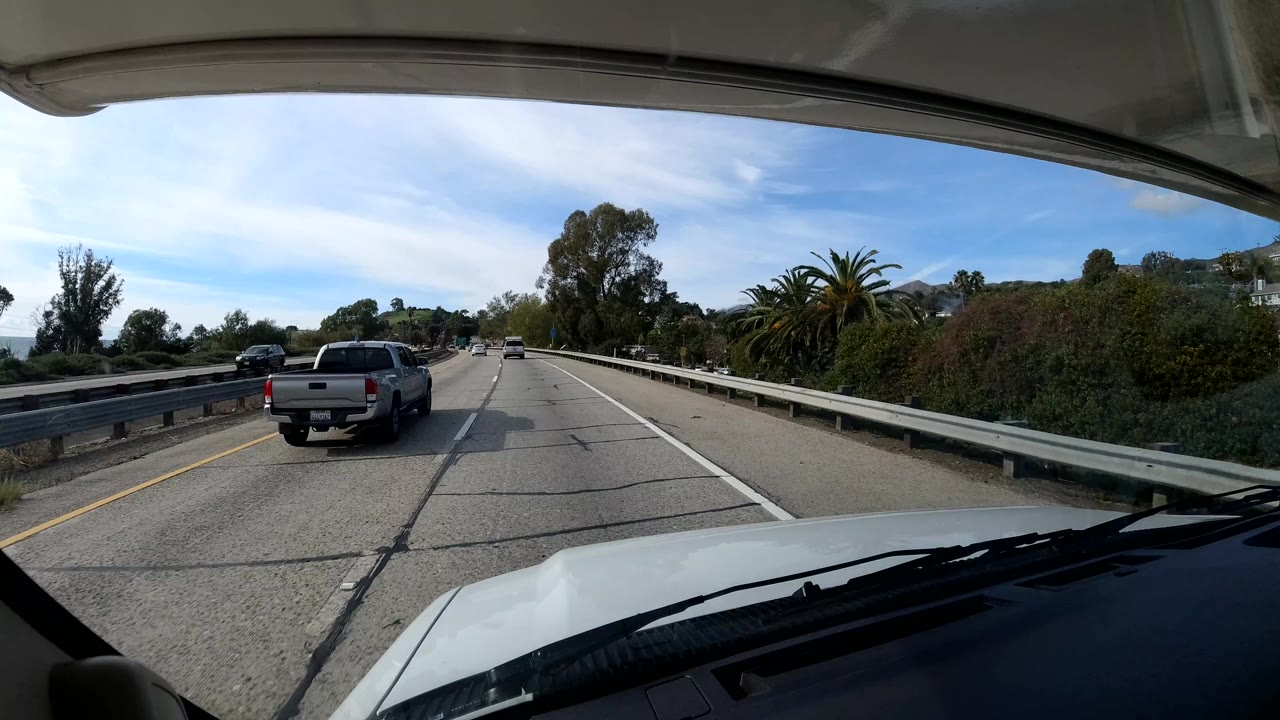 Vehicle-mounted coastal California highway view with a pickup truck ahead, guardrail at left, trees and palms beyond the road, and no visible people.