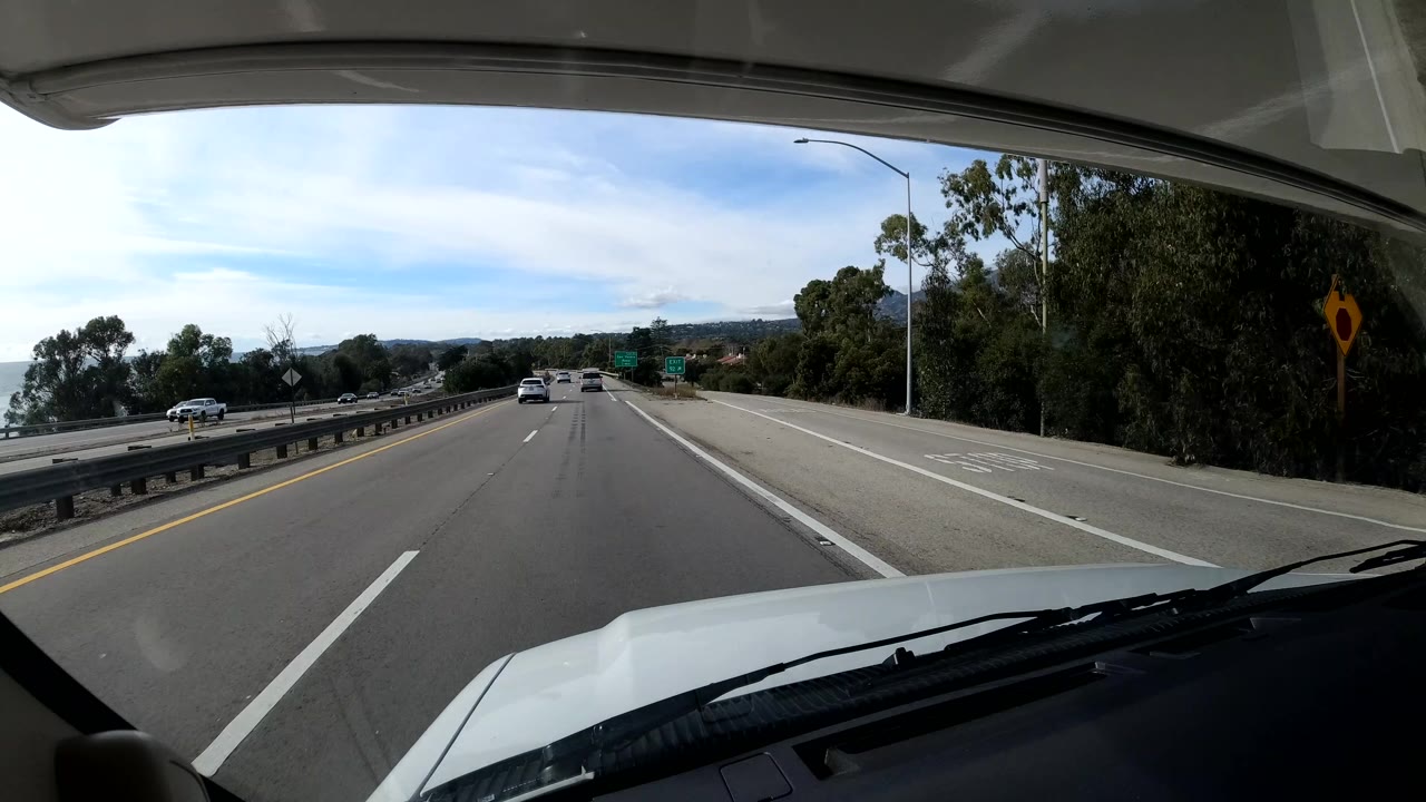 Coastal California highway frame with cars spaced ahead, multiple lanes, guardrails, dense roadside trees, a distant green sign, and no visible people.