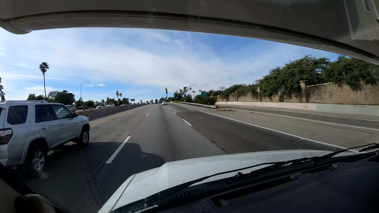 Forward windshield view on a California coast highway with the vehicle hood at bottom, a white SUV in the left lane, shrubs along the right side, and no visible people.