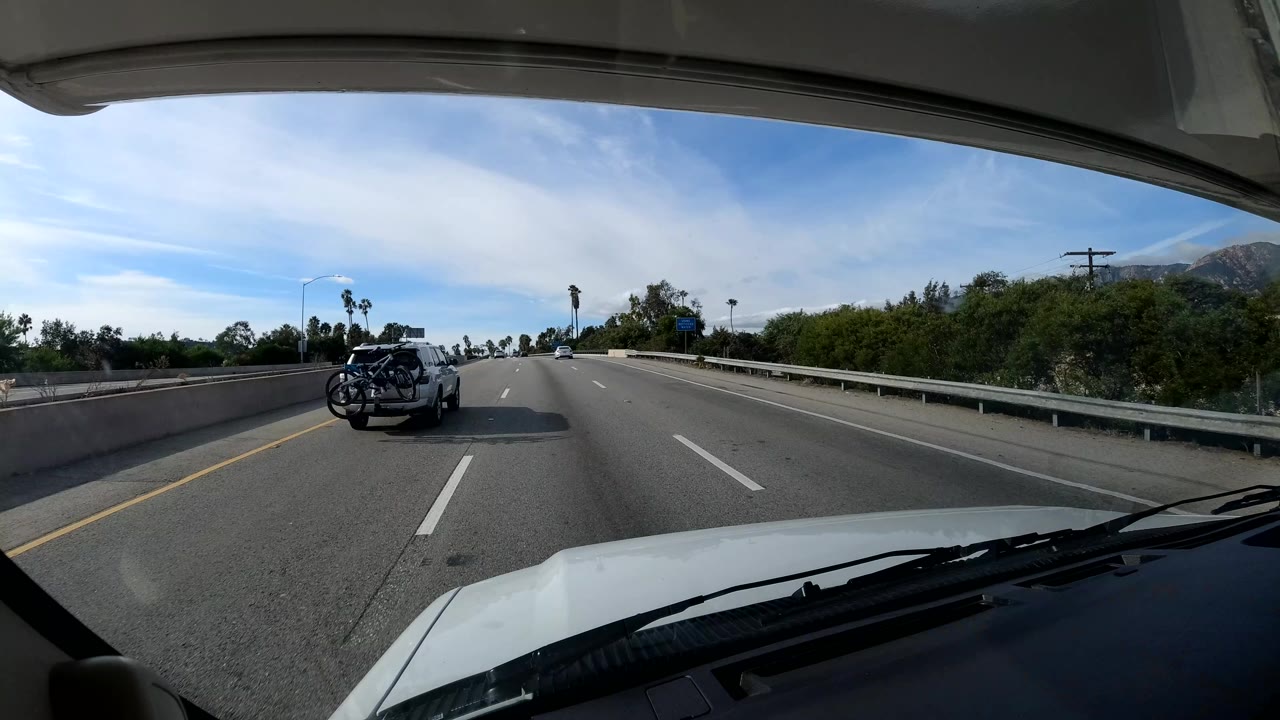 California coast highway view from a moving vehicle, with a car carrying bicycles ahead, guardrail and greenery on the right, distant hills, and no visible people.
